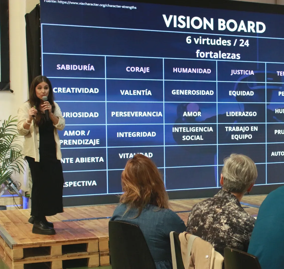 Una presentadora habla al micrófono en un escenario, ante una pantalla con las seis virtudes y las 24 fortalezas del carácter; el público sigue sentado y toma notas.